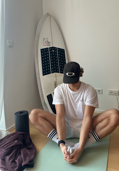 Persona estirándose sobre una esterilla de yoga de color verde claro, vistiendo una camisa blanca, una gorra negra, un reloj negro y calcetines a rayas. Tabla de surf y esterilla de yoga cerca.