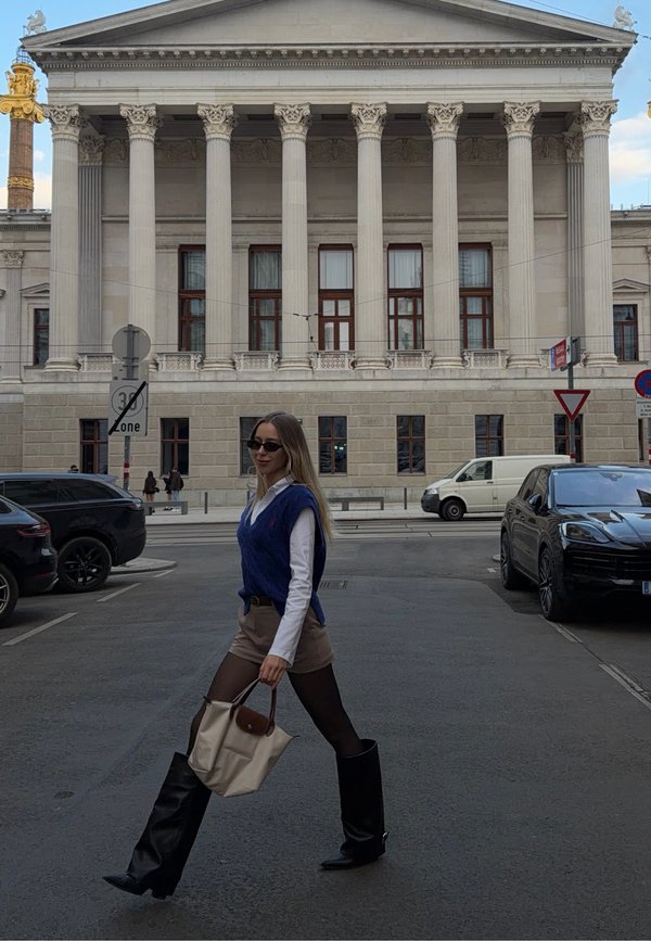 Kobieta w okularach przeciwsłonecznych, niebieskiej kamizelce i czarnych butach przechodząca przez ulicę przed dużym klasycznym budynkiem z kolumnami i zaparkowanymi samochodami.