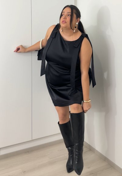 Vestido de satén negro con tirantes anchos y corte holgado, combinado con botas de cuero negro hasta la rodilla, con punta afilada y diseño elegante.