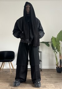 Schwarze Bomberjacke mit Kapuze, oversize Passform, kombiniert mit einem langen schwarzen, gerippten Schal. Locker sitzende schwarze Shorts und klobige schwarze Schuhe.