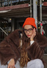 Smeđi kaput od umjetnog krzna s širokim rukavima, narančasta pletena kapa i prozirne naočale; sjedi naslonjen na skelu, s rukom naslonjenom na lice.