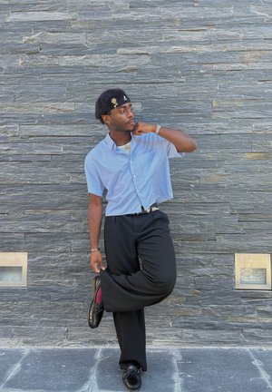 Camisa de rayas azul y blanca de manga corta, pantalones negros, zapatos negros y un sombrero negro con un acento dorado. Fondo de pared de piedra gris.