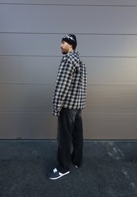 Zwart-wit geruit shirt met een losse fit, gecombineerd met zwarte broek en grijze sneakers met witte accenten. Zwarte beanie met een ontwerp.