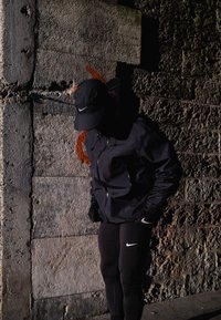 Veste de sport noire et leggings assortis avec un logo Nike blanc. Le modèle porte une casquette noire et se tient devant un mur en pierre texturé.