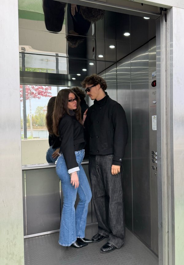 Une femme et un homme vêtus de vêtements sombres et portant des lunettes de soleil se tiennent côte à côte dans un ascenseur ouvert.