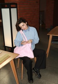 Pullover azul claro com uma camisa de colarinho por baixo, combinado com calças pretas e botas pretas. Segurando uma bolsa de tote rosa felpuda enquanto está sentado.