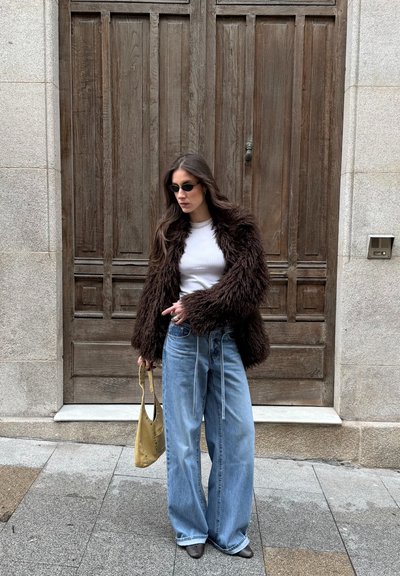 Mujer con chaqueta de piel marrón oscuro, camiseta blanca, vaqueros de pierna ancha y gafas de sol, sosteniendo un bolso amarillo, de pie frente a una puerta de madera.