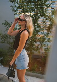 Black sleeveless top, high-waisted light blue denim shorts with frayed hem, black handbag, and sunglasses, set against greenery.