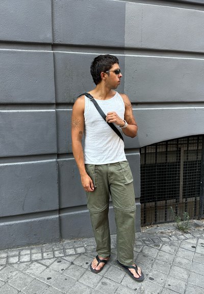 Camiseta sin mangas blanca, pantalones cargo verde oliva con bolsillos, chanclas negras y una bolsa de hombro negra. Cabello corto y oscuro, gafas de sol puestas.
