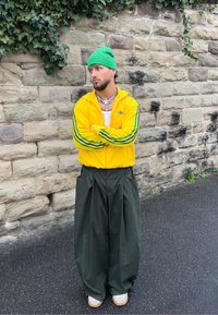 Veste zippée jaune Adidas avec des rayures vertes, pantalons larges noirs, bonnet vert, et baskets blanches. Positionné contre un mur en pierre.
