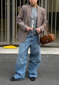 Osoba v nadrozmernom hnedo károvanom saku, sivom gombíkovom kardigáne, širokých modrých džínsach, čiernych topánkach s rozdelenými špičkami, drží hnedú koženú kabelku.