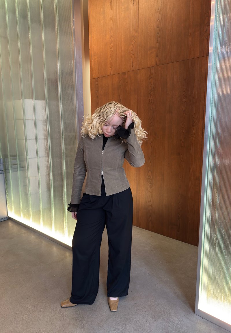 Lichtbruine corduroy jas met rits, gecombineerd met zwarte wijde broek en beige platte schoenen, tegen een achtergrond van hout en gestructureerd glas.