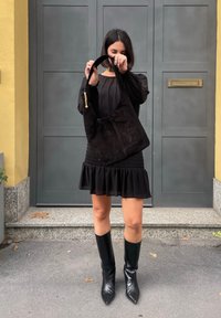 Μαύρη σουέντ τσάντα ώμου με καμπύλη λαβή και διακοσμητική χρυσή λεπτομέρεια. Φορεμένη από ένα άτομο σε μαύρο φόρεμα και μαύρες μπότες μέχρι το γόνατο.