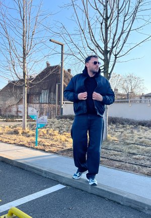 Chaqueta bomber negra, sudadera negra y pantalones azul marino. Zapatillas gris, gafas de sol, y de pie en una acera cerca de árboles desnudos y edificios.