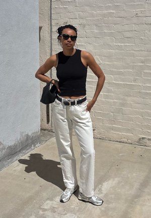 Mujer con top negro sin mangas, pantalones blancos, gafas de sol, sosteniendo bolso negro, de pie junto a una pared de ladrillo clara bajo la luz del sol.