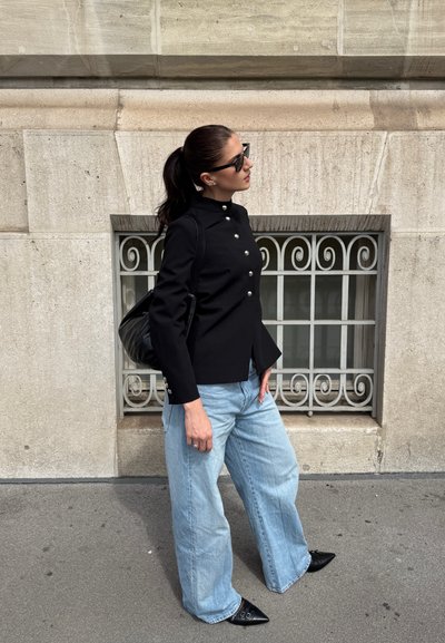 Chaqueta negra con cuello, detalles de botones plateados, combinada con jeans de pierna ancha de color azul claro y zapatos negros de punta, contra un muro de piedra beige.
