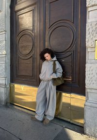 Person med krøllet hår iført grå hættetrøje og posede grå bukser, lænende sig op ad store udsmykkede mørke trødøre med guldfarvede paneler i bunden.