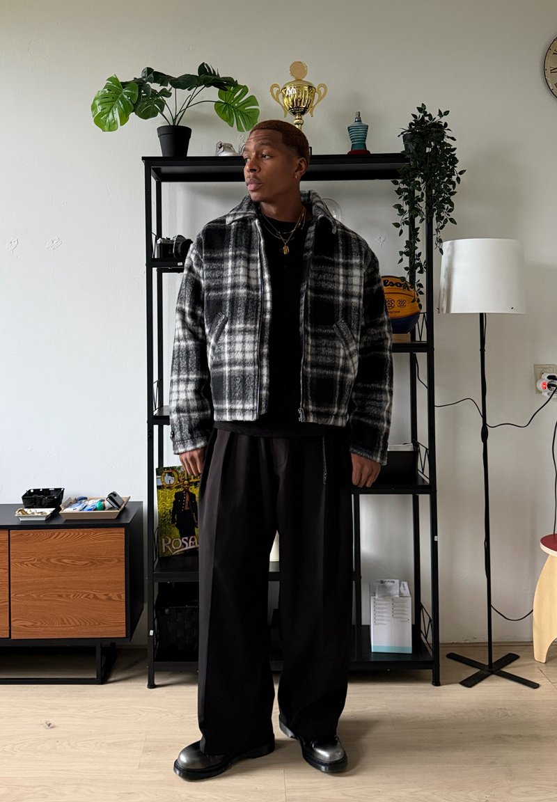 Veste à fermeture éclair à carreaux noire et blanche en polaire, associée à un pantalon large noir et des bottes noires. En arrière-plan, des plantes et de la décoration.