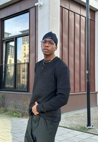 Jovem de camisa de manga longa preta e calças, usando óculos e um gorro preto, em pé na calçada urbana perto de um edifício com grandes janelas.