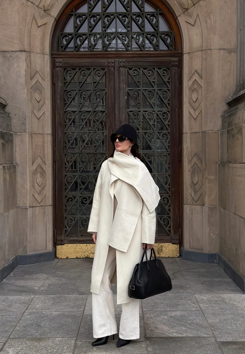 White wool coat with an asymmetrical drape, flared white trousers, black heeled boots, black handbag, and wide-brimmed black hat.