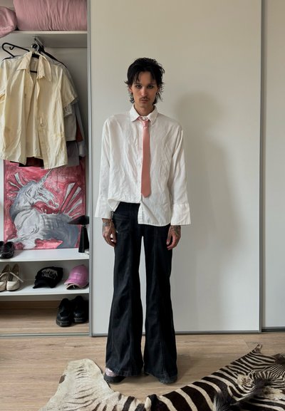 Persona con cabello oscuro y tatuajes lleva una camisa blanca, una corbata rosa y pantalones acampanados negros, de pie en una habitación con un armario y una alfombra de cebra.