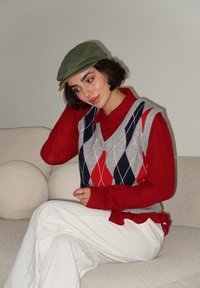 Γκρι γιλέκο με διαμαντόσχημα σχέδια σε κόκκινο, ναυτικό μπλε και λευκό πάνω από κόκκινο πουκάμισο με γιακά. Λευκά παντελόνια με φαρδιά πόδια και ελιά πράσινο καπελάκι.