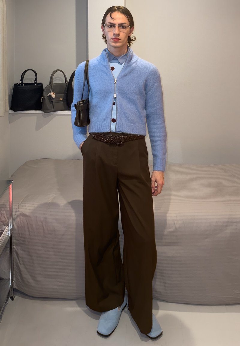 Personne portant un cardigan court bleu clair, une chemise bleue, un pantalon marron à jambes larges et des chaussures bleu clair, debout dans une pièce avec des sacs à main sur une étagère.
