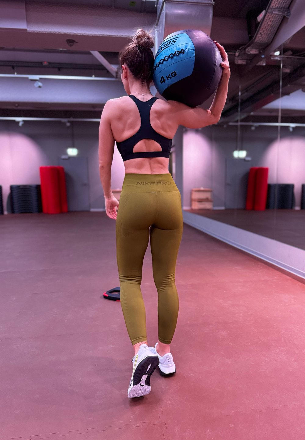Femme en brassière de sport noire et legging vert tenant un ballon de médecine de 4 kg sur son épaule dans une salle de sport avec un mur miroir et des tapis d'exercice.