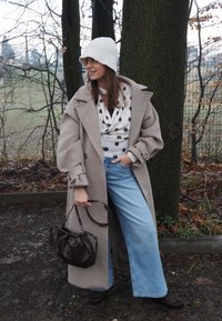 Manteau oversize en mélange de laine beige, haut blanc à pois, jean à jambes larges, sac seau marron, bonnet blanc en tricot, dans un cadre boisé en plein air.