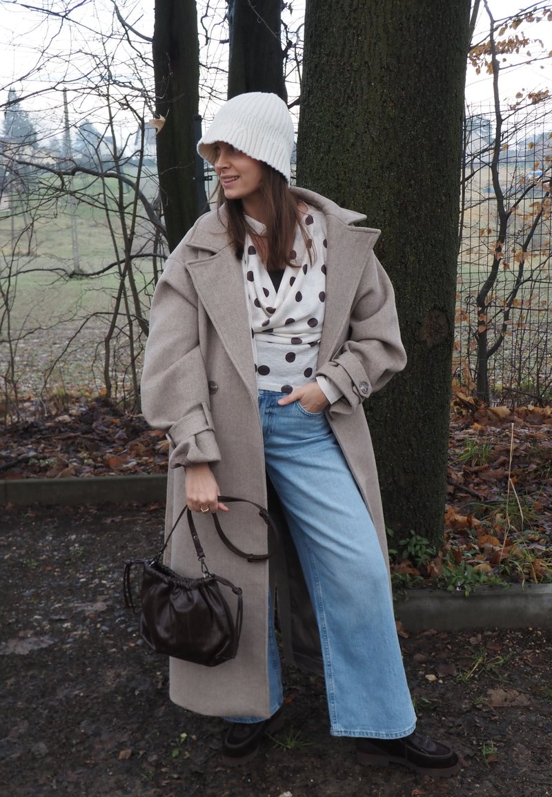 Manteau oversize en mélange de laine beige, haut blanc à pois, jean à jambes larges, sac seau marron, bonnet blanc en tricot, dans un cadre boisé en plein air.