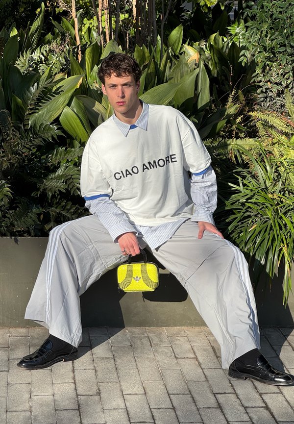 T-shirt bianca oversize con il testo "CIAO AMORE", indossata sopra una camicia button-up azzurro chiaro. Pantaloni grigi larghi, scarpe nere e una borsa gialla piccola.