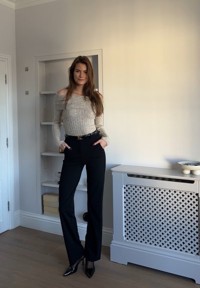 Suéter color beige con los hombros al descubierto, pantalones negros a medida y tacones negros puntiagudos. Los pantalones tienen pasadores para cinturón y un acabado suave. Entorno interior sencillo.