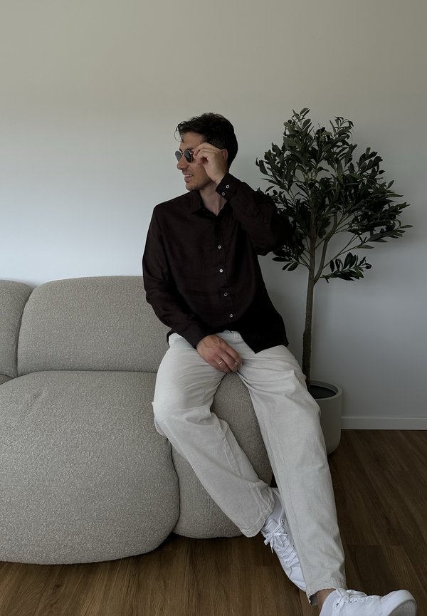 Mies, jolla aurinkolasit, tumma paita ja vaaleat housut, istuu beigeillä sohvalla, jonka vieressä on ruukkukasvi puulattialla.
