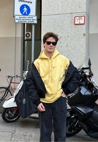 Oseba v rumeni kapuci, ki je oblečena pod črno puhovko, v temnih hlačah in sončnih očalih, stoji poleg rdečega kolesa in motornega kolesa.