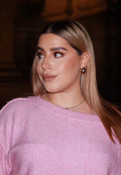 Suéter rosa claro de corte holgado y con cuello acanalado, combinado con grandes pendientes de aro dorados y una delicada cadena. Maquillaje en tonos neutros.