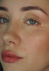 Nærbilde av et ansikt med grønnblå øyne, en hjertformet gull septumring og glansfulle, myke rosa lepper. Glitrende øyesminke.
