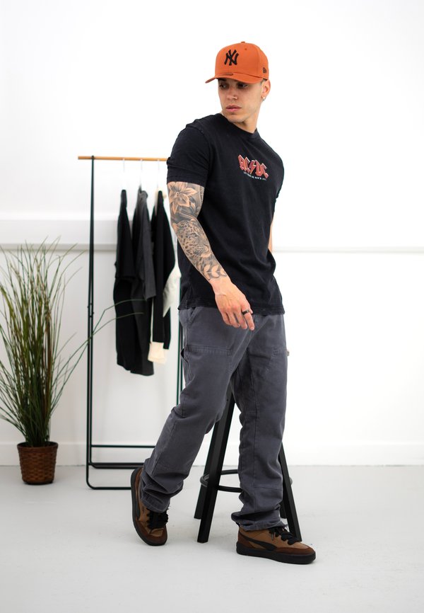 Schwarzes T-Shirt mit rographic, graue Cargo-Hosen, braune und schwarze Sneaker, orangefarbene Mütze, tätowierter Arm, steht neben einer Kleiderstange und einer Pflanze.