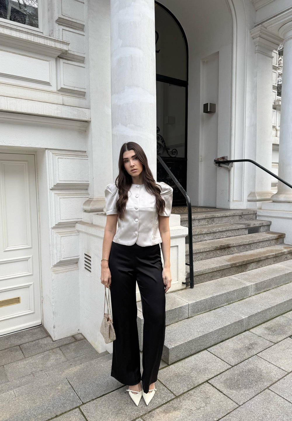 Haut en satin beige clair à manches bouffantes avec boutons, associé à un pantalon noir fluide et des chaussures blanches pointues. Tient un sac à main beige texturé.