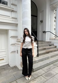 Haut en satin beige clair à manches bouffantes avec boutons, associé à un pantalon noir fluide et des chaussures blanches pointues. Tient un sac à main beige texturé.