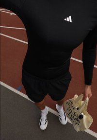 Camisola desportiva de mangas longas preta com logo branco, calções pretos, sapatilhas brancas, e uma mala de mão creme com texto verde, num pista de corrida.