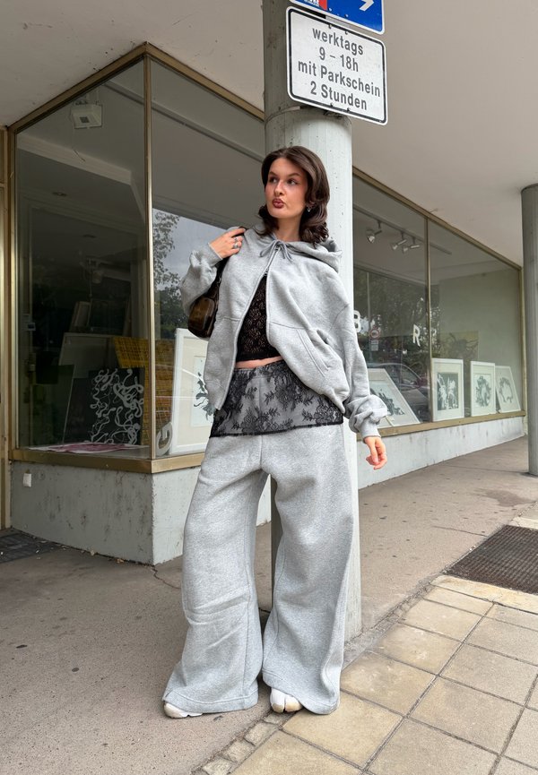 Young woman wearing an oversized grey hoodie and wide pants stands next to a street sign and building with large glass windows and artwork inside.