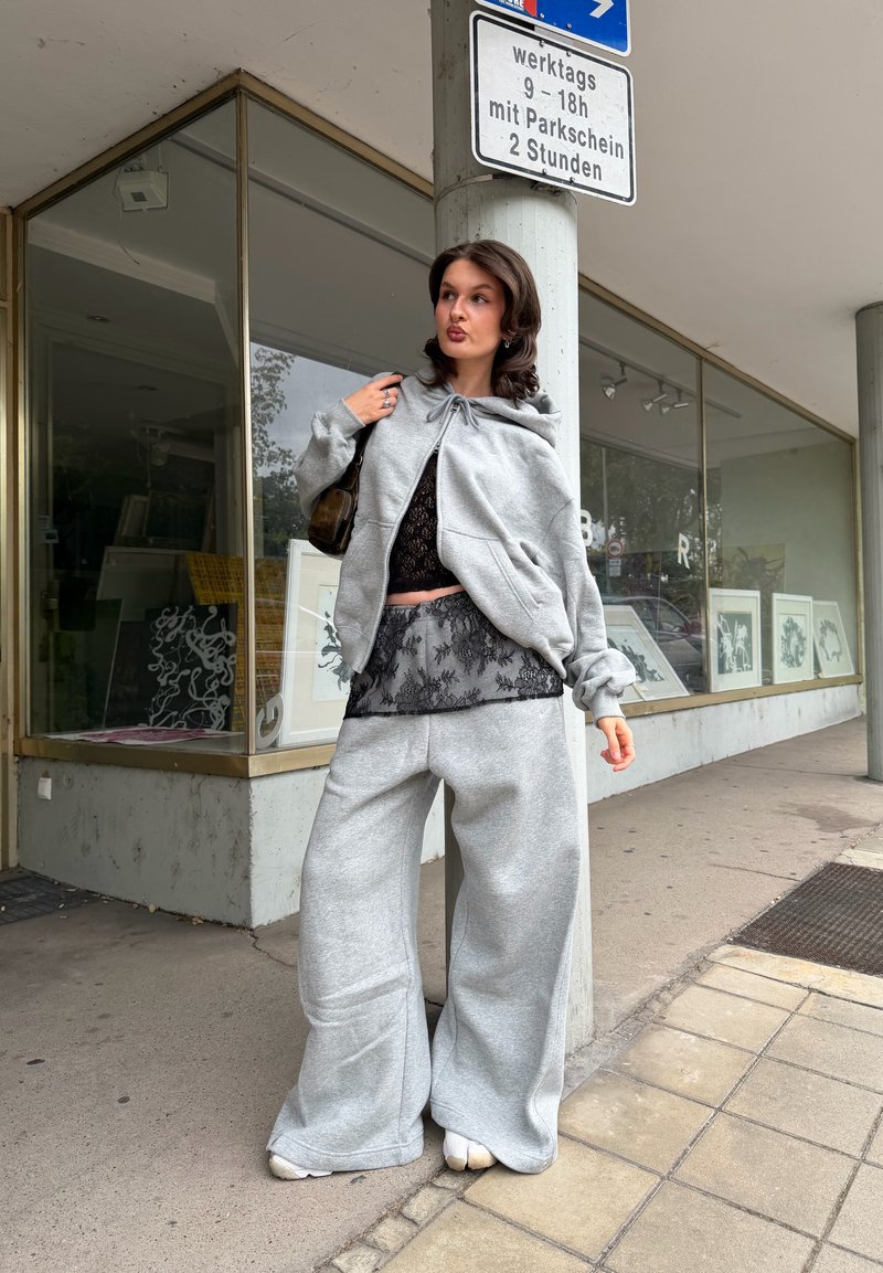 Joven mujer con sudadera gris de gran tamaño y pantalones anchos está junto a una señal de la calle y un edificio con grandes ventanales de vidrio y obras de arte en su interior.
