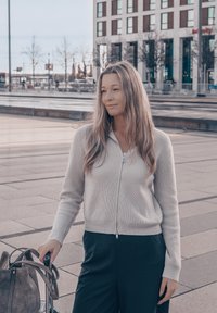 Cardigan gris clair en maille côtelée avec fermeture éclair, associé à un pantalon foncé. Un sac gris avec des bretelles est posé sur une poignée de bagage. Arrière-plan d'une place urbaine.