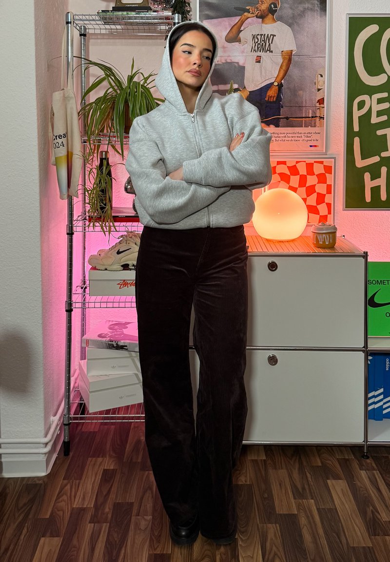Casaco com capuz cinza claro e fecho zip, combinado com calças pretas de bombazina de cintura alta. Em pé sobre um chão de madeira com objetos coloridos ao fundo.