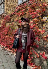 Nainen viininpunaiseen nahkatakkiin, hameeseen, lippalakkiin ja mustiin sukkahousuihin pukeutuneena seisoo seinää vasten, joka on peittynyt punaiseen ja keltaiseen syksyn köynnösruusuun.