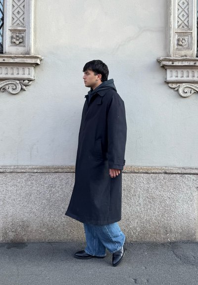 Joven con un abrigo largo negro, vaqueros azules y zapatos negros caminando junto a una pared de piedra y estuco texturizada con detalles arquitectónicos decorativos.