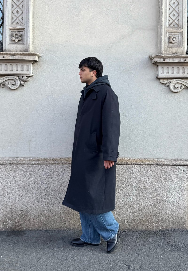 Jeune homme en long manteau noir, jean bleu et chaussures noires marchant à côté d'un mur en pierre texturé et stuc avec des détails architecturaux décoratifs.