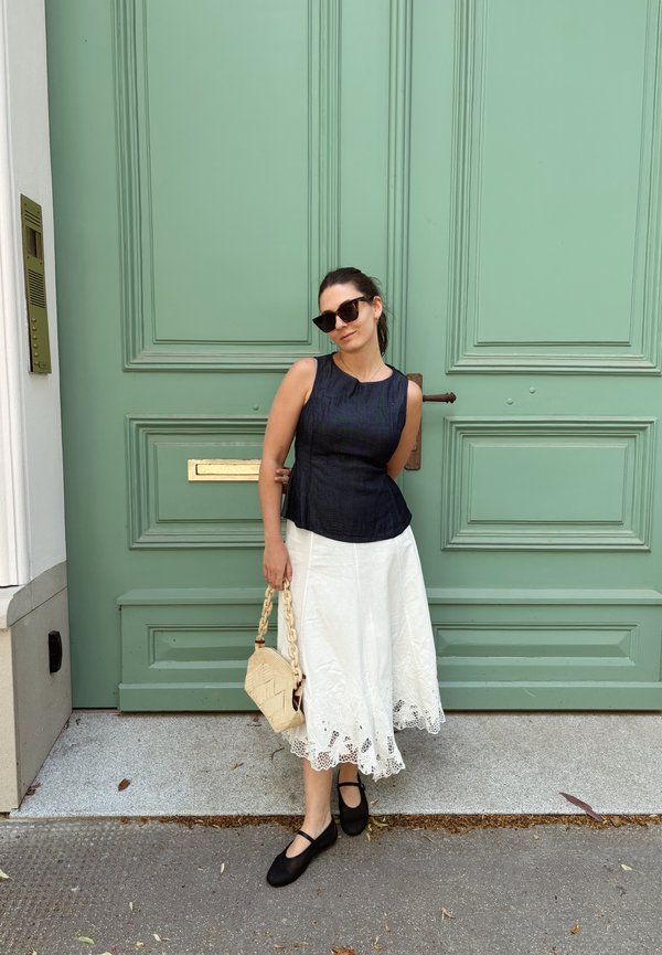 Frau mit Sonnenbrille, ärmellosem schwarzem Oberteil, weißem Rock, schwarzen Schuhen, hält eine geflochtene Handtasche und steht vor einer großen grünen Tür.