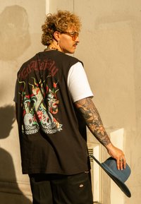 Schwarzes ärmelloses Hemd mit einem bunten Totenkopf- und Schlangendesign auf dem Rücken, getragen über einem weißen T-Shirt, mit beigen Hosen und einem blauen Hut.