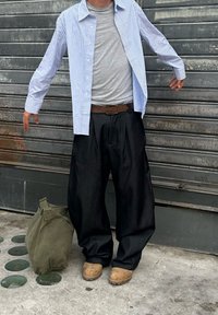 Dryžuota ilgomis rankovėmis marškiniai, pilkas marškinėlis, juodos plačios kelnės, rudas dirželis ir rudi mediniai šlepetės. Žalias kanapių maišelis padėtas ant žemės šalia.